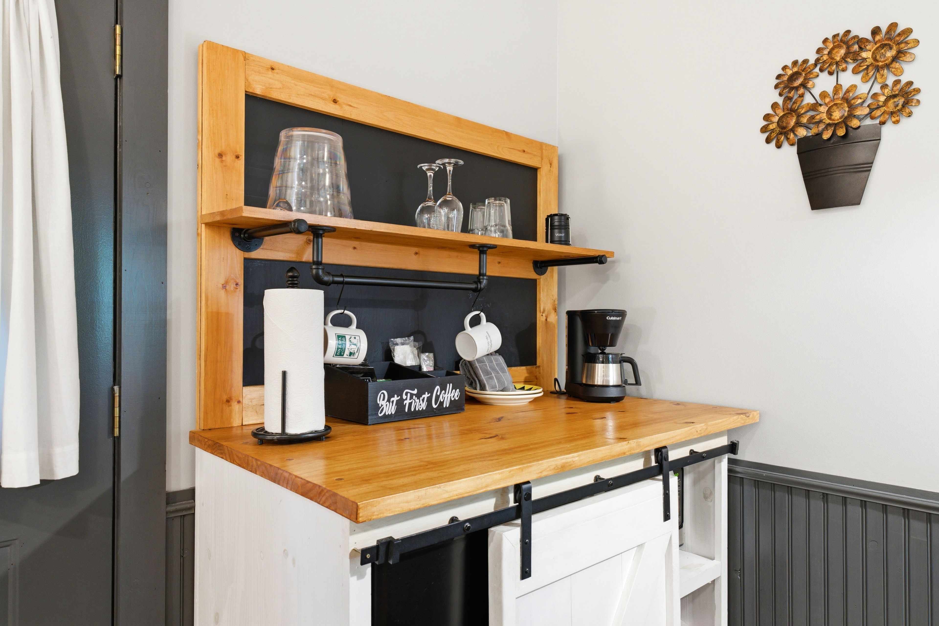 En køkkenbordplade med kaffemaskine, en hylde med drikkeglas og et skilt med teksten 'But First Coffee'.