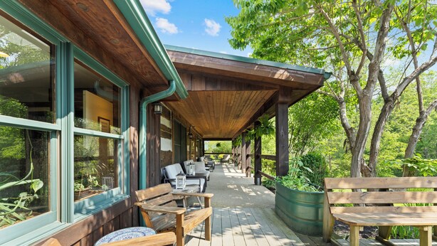 A covered outdoor seating area with wooden benches and tables, surrounded by greenery and trees.