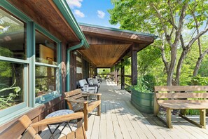 A covered outdoor seating area with wooden benches and tables, surrounded by greenery and trees.