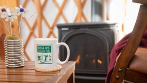 A white mug with "INN & SPA" and "Cedar Falls" logo, a metal vase with artificial flowers, and a wooden table in a cozy room with a fireplace.