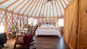 A yurt interior with a bed, rocking chair, and a small table with a lamp.