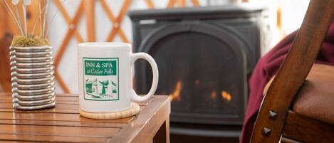 A white mug with "INN & SPA" and "Cedar Falls" logo, a metal vase with artificial flowers, and a wooden table in a cozy room with a fireplace.