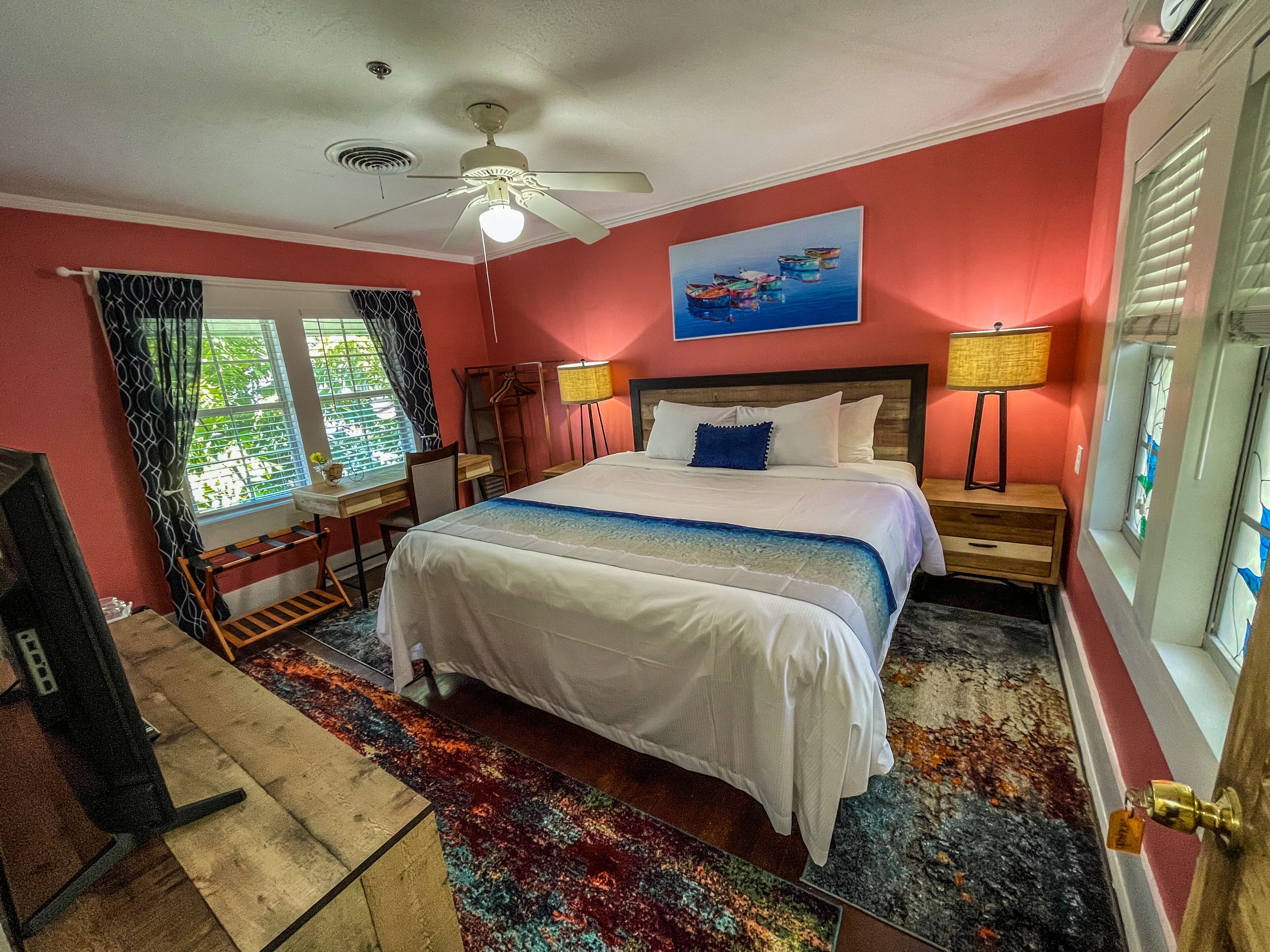 A bedroom with a bed, a ceiling fan, a window with curtains, a painting on the wall, and a nightstand with a lamp.