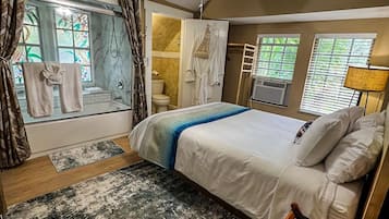 A bedroom with a bed, a ceiling fan, a window with curtains, a bathroom with a shower, and a view of the outside.