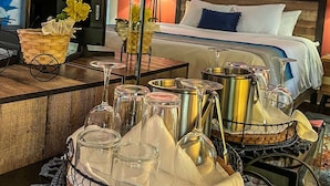 Une chambre d’hôtel avec un lit, un ventilateur de plafond, un tableau représentant des bateaux, une table avec des verres et un panier de fleurs.