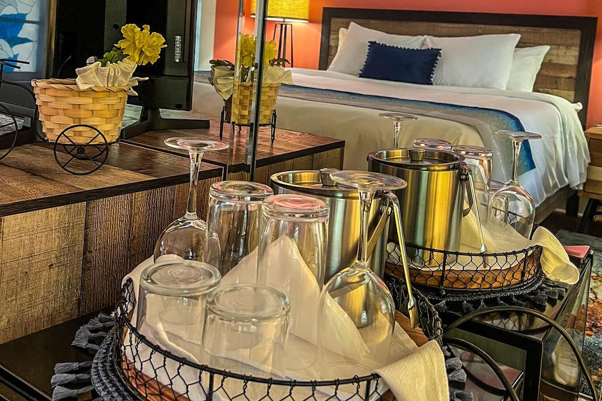 Une chambre d’hôtel avec un lit, un ventilateur de plafond, un tableau représentant des bateaux, une table avec des verres et un panier de fleurs.