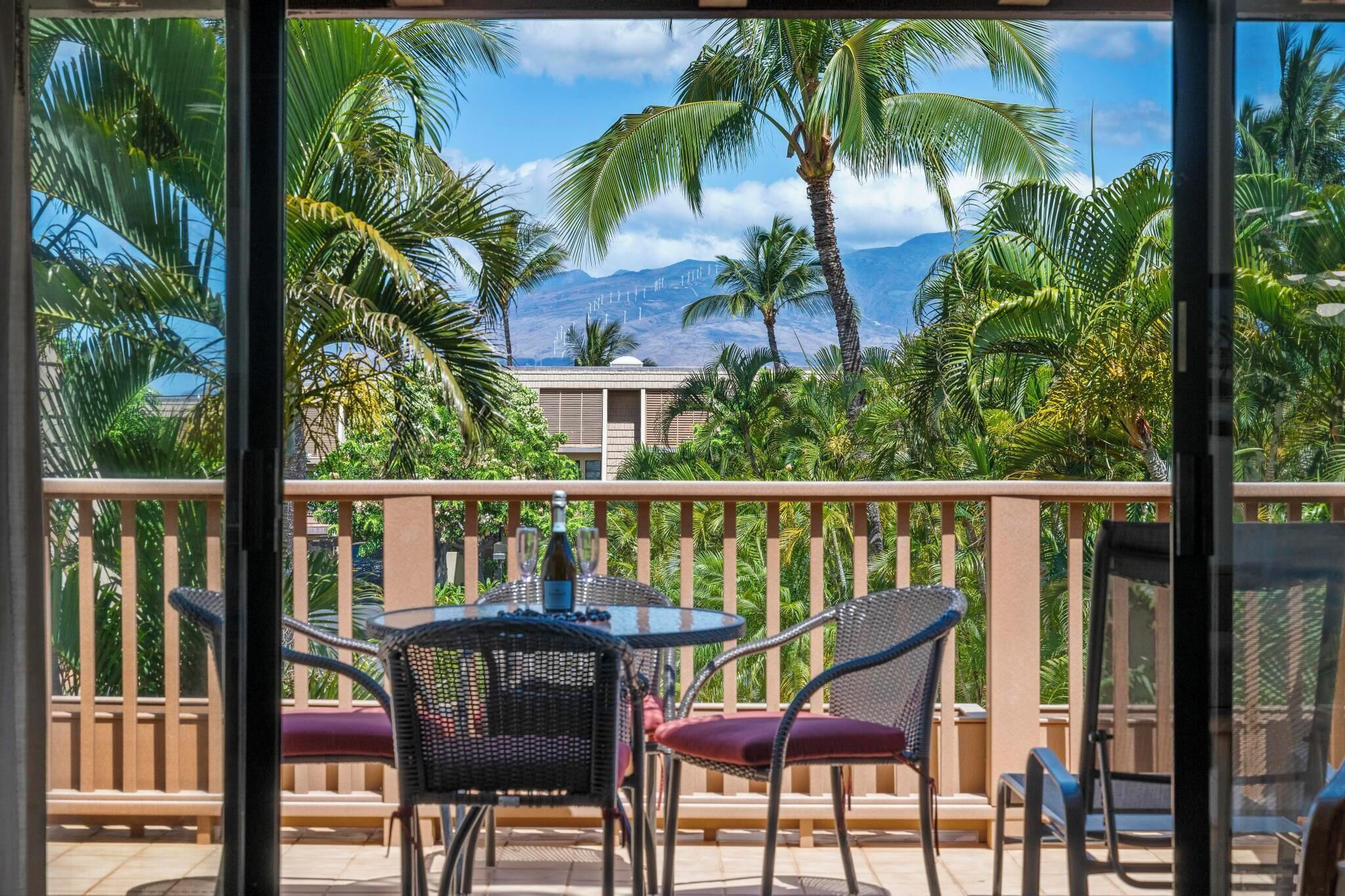 A balcony with a table and chairs, overlooking a tropical landscape with palm trees and a mountain in the background.