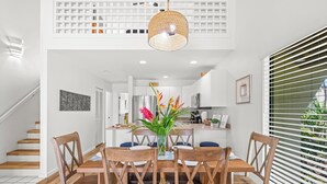 Un comedor con una mesa y sillas de madera, una lámpara colgante, y vistas a la cocina y la escalera al fondo.