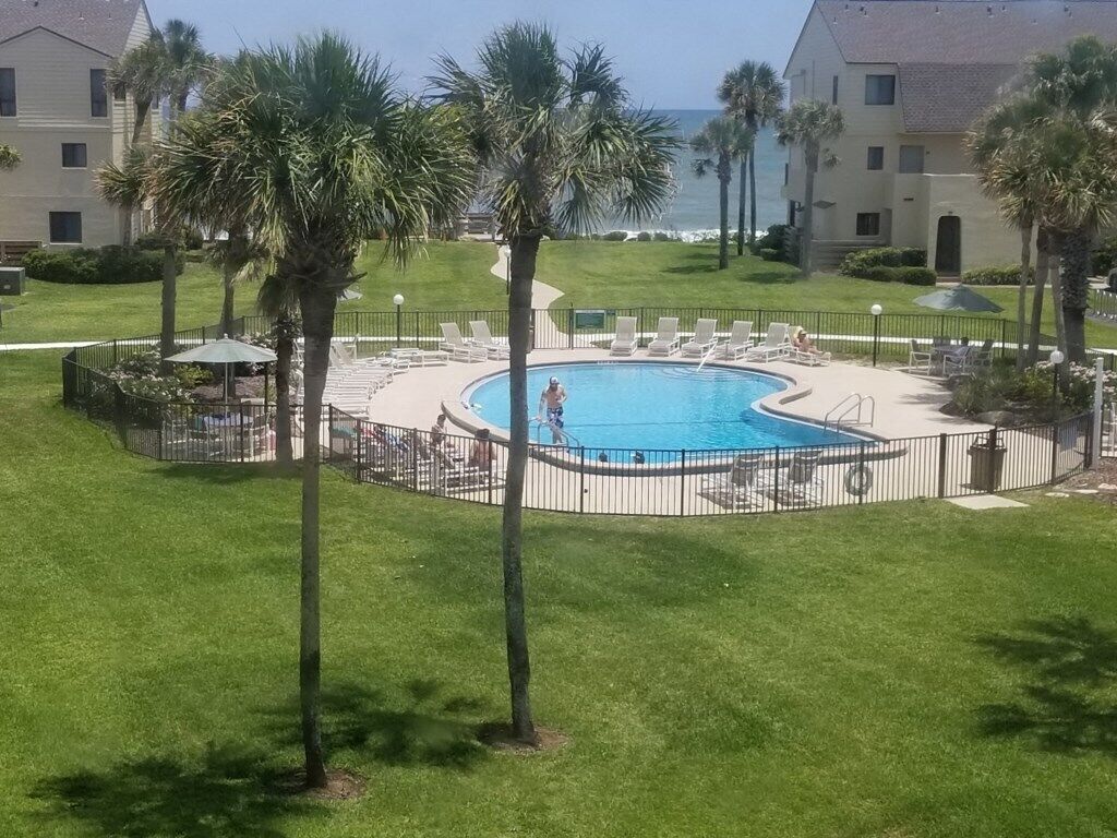 A swimming pool surrounded by lounge chairs and palm trees, with a view of the ocean and buildings in the background.