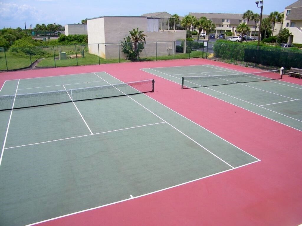 Un court de tennis extérieur comprenant plusieurs terrains, entouré d’une clôture et de verdure.