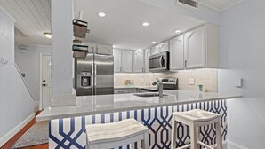 A modern kitchen with white cabinetry, stainless steel appliances, and a central island with bar stools. - Ocean Club 37 (St. Augustine)