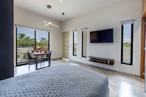 A modern bedroom with a large bed, a dining area with a table and chairs, a TV mounted on the wall, and a sliding glass door leading to a balcony with a view of palm trees. - El Desierto by Naya Homes (San José del Cabo)