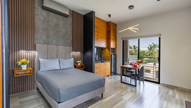 A modern bedroom with a large bed, wooden paneling, a built-in kitchen, and a balcony with a view. - El Desierto by Naya Homes (San José del Cabo)