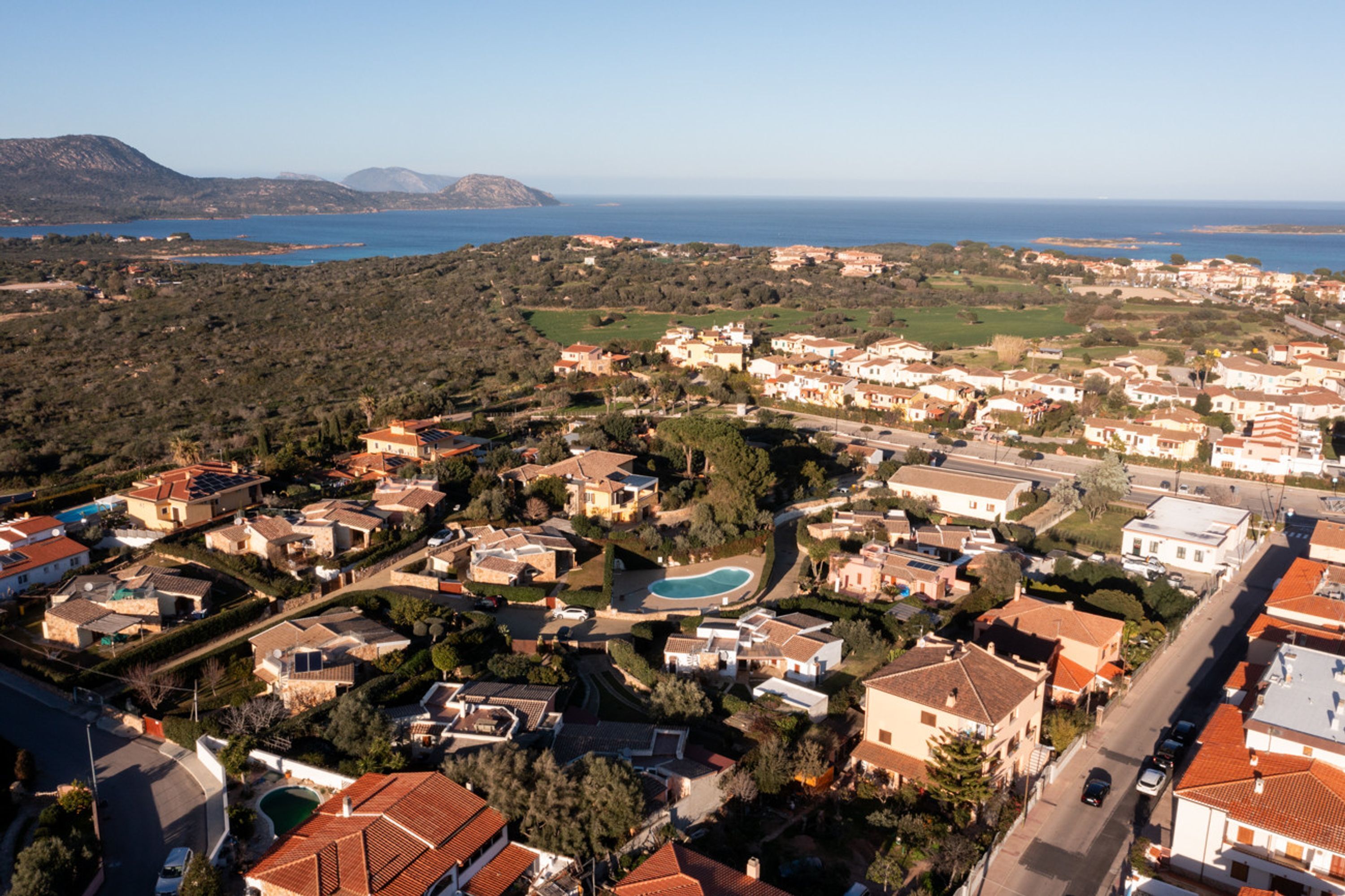 An aerial view of a coastal residential area with houses, a swimming pool, and a clear view of the sea and mountains in the background.