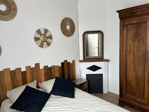 A bedroom with a wooden headboard, a bed with white and dark blue bedding, a mirror above a fireplace, and a wooden wardrobe. - Maison de Ville Avec Jardin Proche du Pont Canal (Briare)