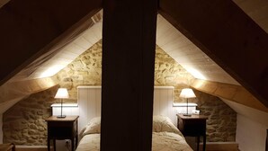 An attic bedroom with a stone wall, a bed with a floral duvet, two bedside tables with lamps, and a wooden chair. - House, Central Village location, near cafe/boulangerie, 300m from coast. (Saint Armel)