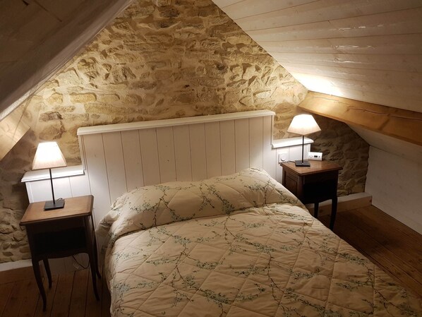 A bedroom with a stone wall, a wooden ceiling, a bed with a floral quilt, and two bedside lamps.
