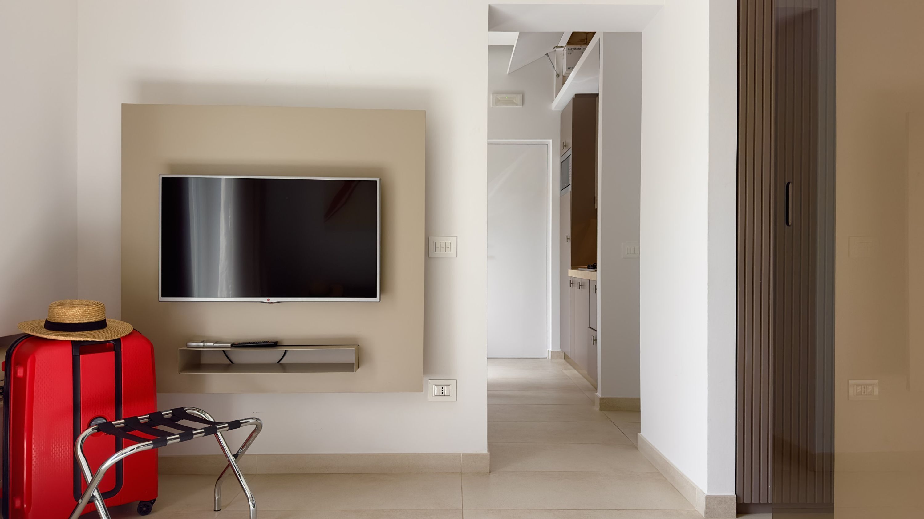 A modern hotel room with a flat-screen TV mounted on the wall, a red suitcase, and a trolley.