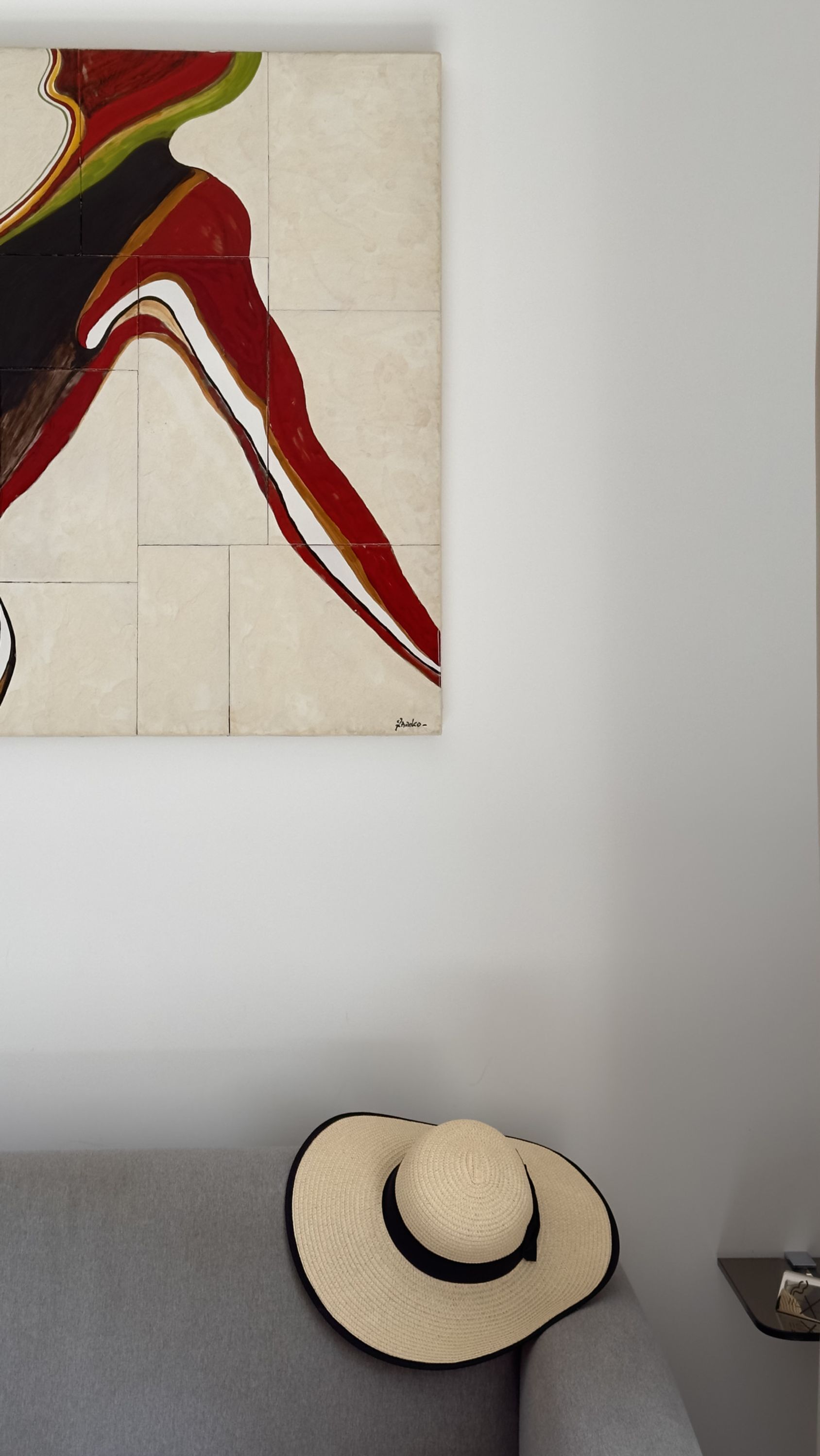 A room with a large abstract painting on the wall and a straw hat on a grey sofa.