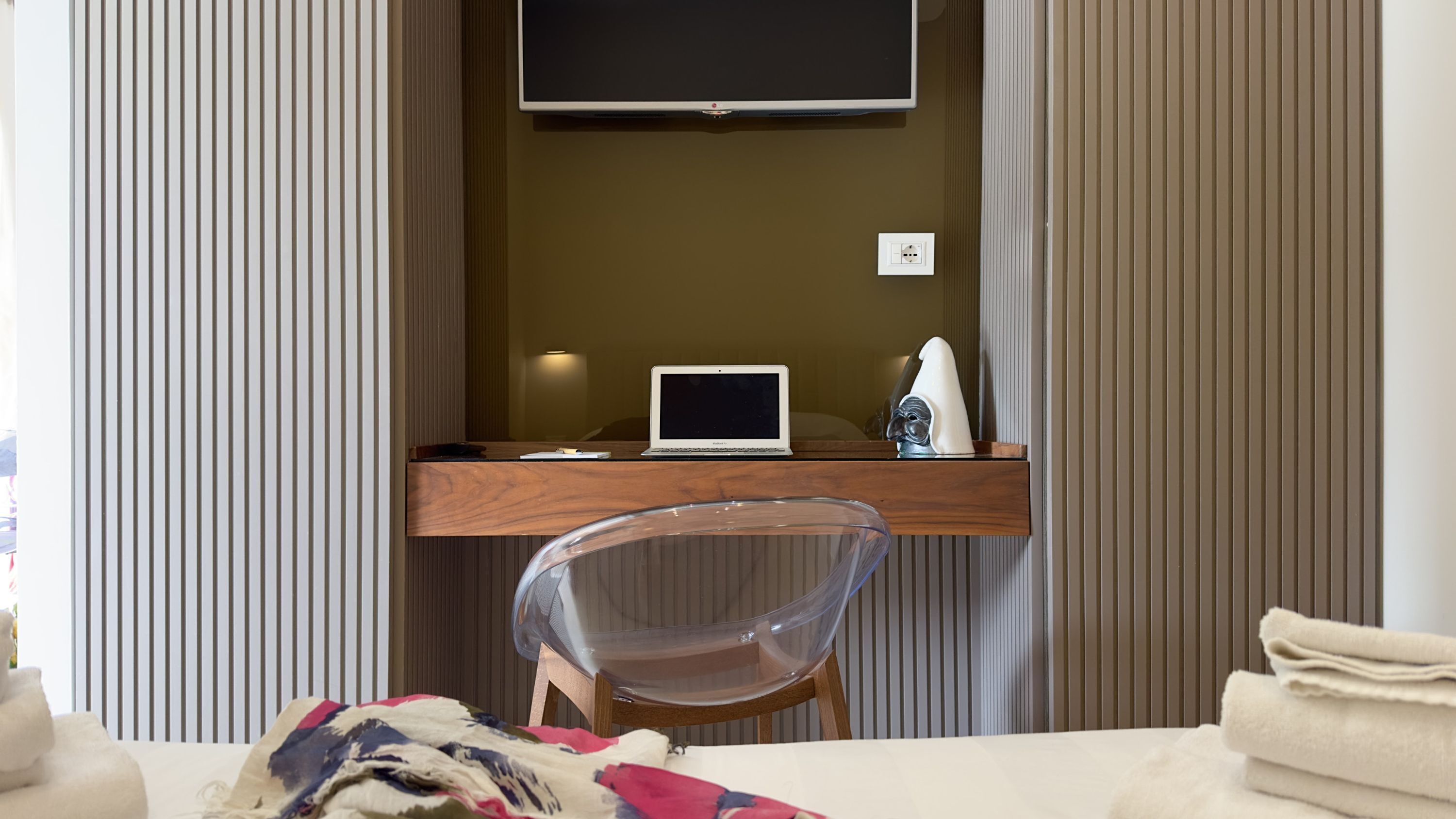 Une chambre d’hôtel avec un bureau en bois, un ordinateur portable, une chaise transparente, un lit recouvert d’une couverture colorée et un téléviseur fixé au mur.