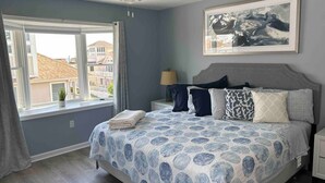 Ein Schlafzimmer mit Bett, Nachttisch, Lampe, Deckenventilator, einem Fenster mit Vorhängen und einem gerahmten Bild an der Wand.