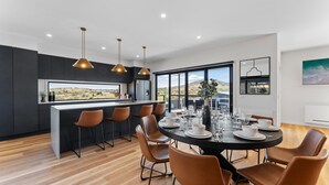 A modern kitchen with a central island, large windows offering a view of the countryside, and a dining area with a round table set for six. - 1B Alpine View Retreat - 4BR Luxurious Escape l Mountain Views l Fast WiFi (Jindabyne)