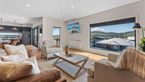 A spacious living area with a large window offering a view of the countryside. There is a dining area with a table and chairs, and a kitchen visible through an open door. - 1B Alpine View Retreat - 4BR Luxurious Escape l Mountain Views l Fast WiFi (Jindabyne)