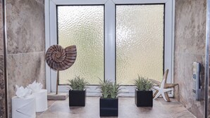 Une salle de bain avec un sol en marbre, une grande fenêtre en verre dépoli, trois plantes en pot, une décoration en forme d’étoile de mer, une chaise en bois et un distributeur de mouchoirs.