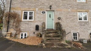 Une petite maison en pierre avec une porte verte, un petit porche et un chemin de gravier menant à l’entrée.