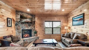 A cozy cabin interior with a stone fireplace, leather sofas, and a large window. - New! Pet Friendly, Private Pool, Media Room, Golf! (Sevierville)