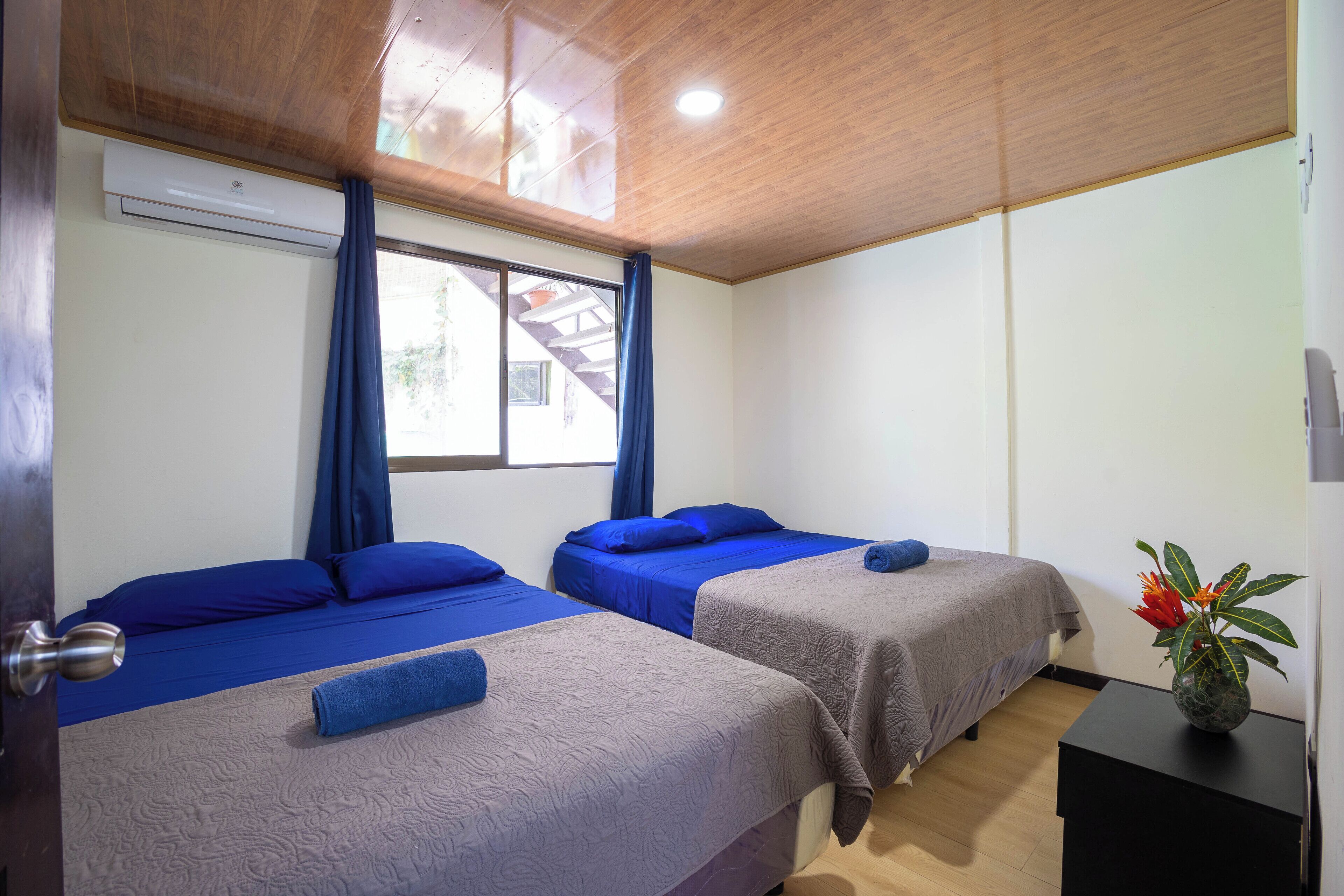 A room with two beds, a window with curtains, an air conditioner, and a vase with flowers on a nightstand.