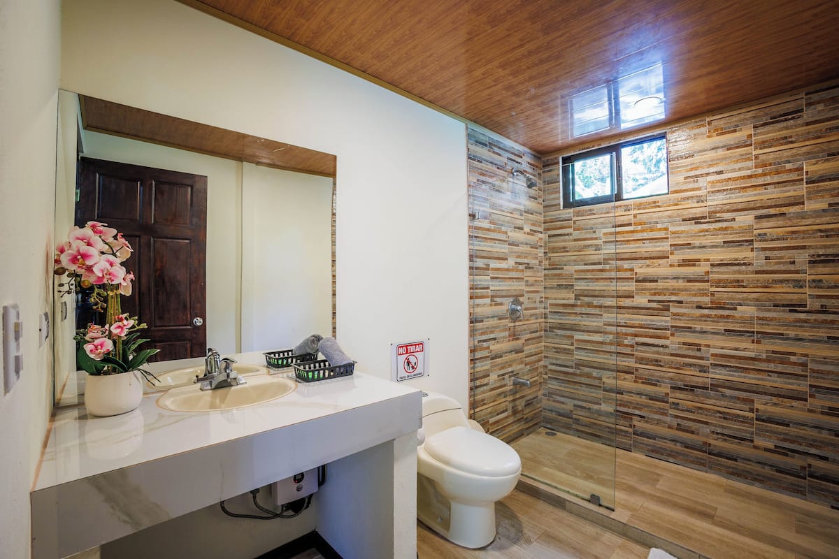 A bathroom with a wooden ceiling, a white sink, a toilet, and a shower with a stone wall.