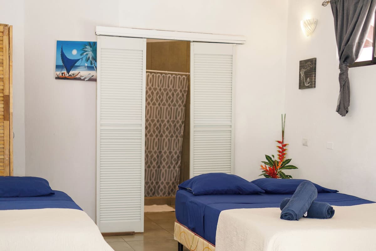 A hotel room with two beds, a window with a curtain, a door with a patterned blind, and a wall with a painting and a plant.