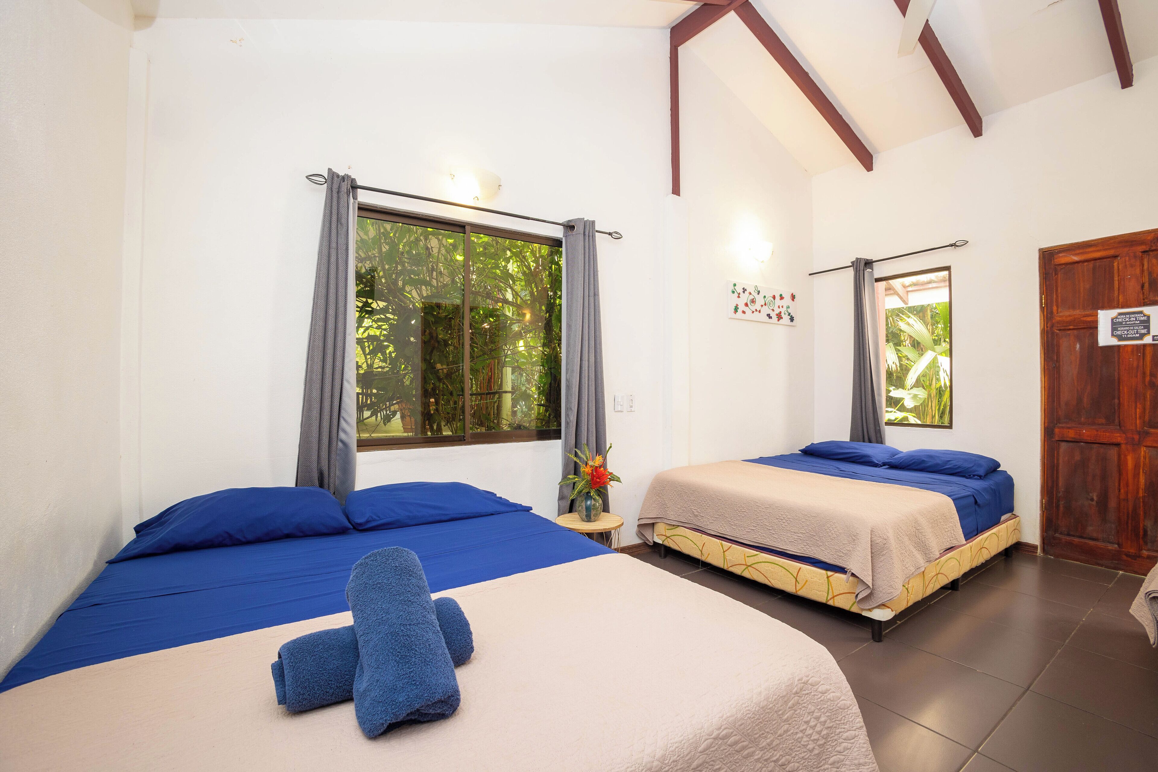 Una habitación con dos camas, cada una con ropa de cama azul, una mesita con un jarrón y una puerta de madera.
