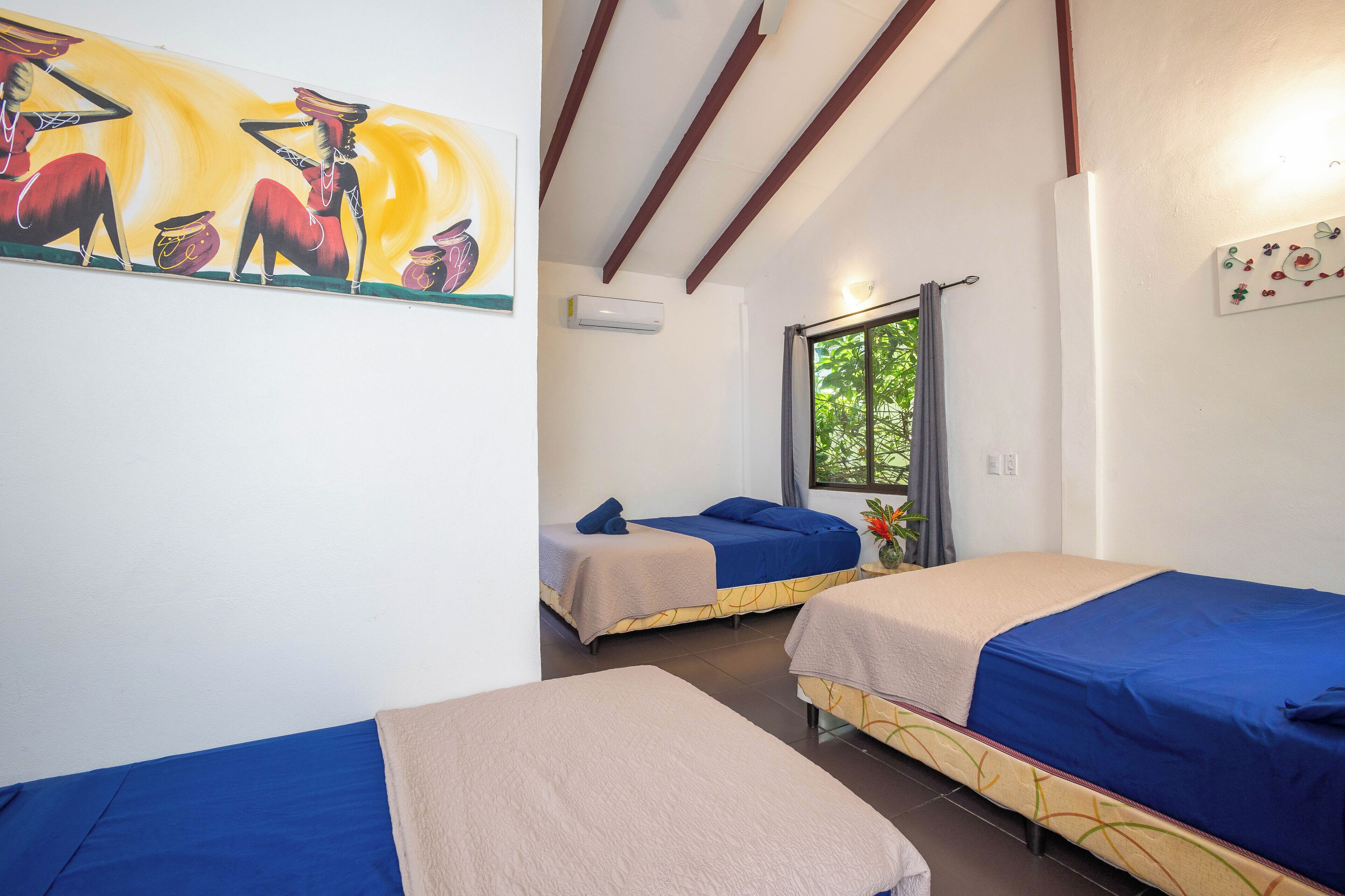 A room with two beds, each with a blue and beige bedspread, a painting on the wall, and a window with a view of greenery.