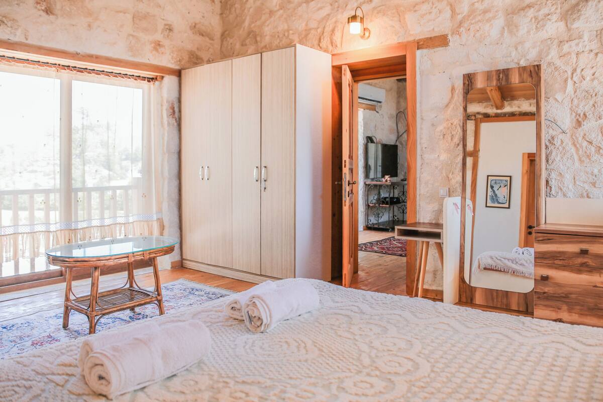 A room with a bed, a glass-top table, a wooden cabinet, and a large window.