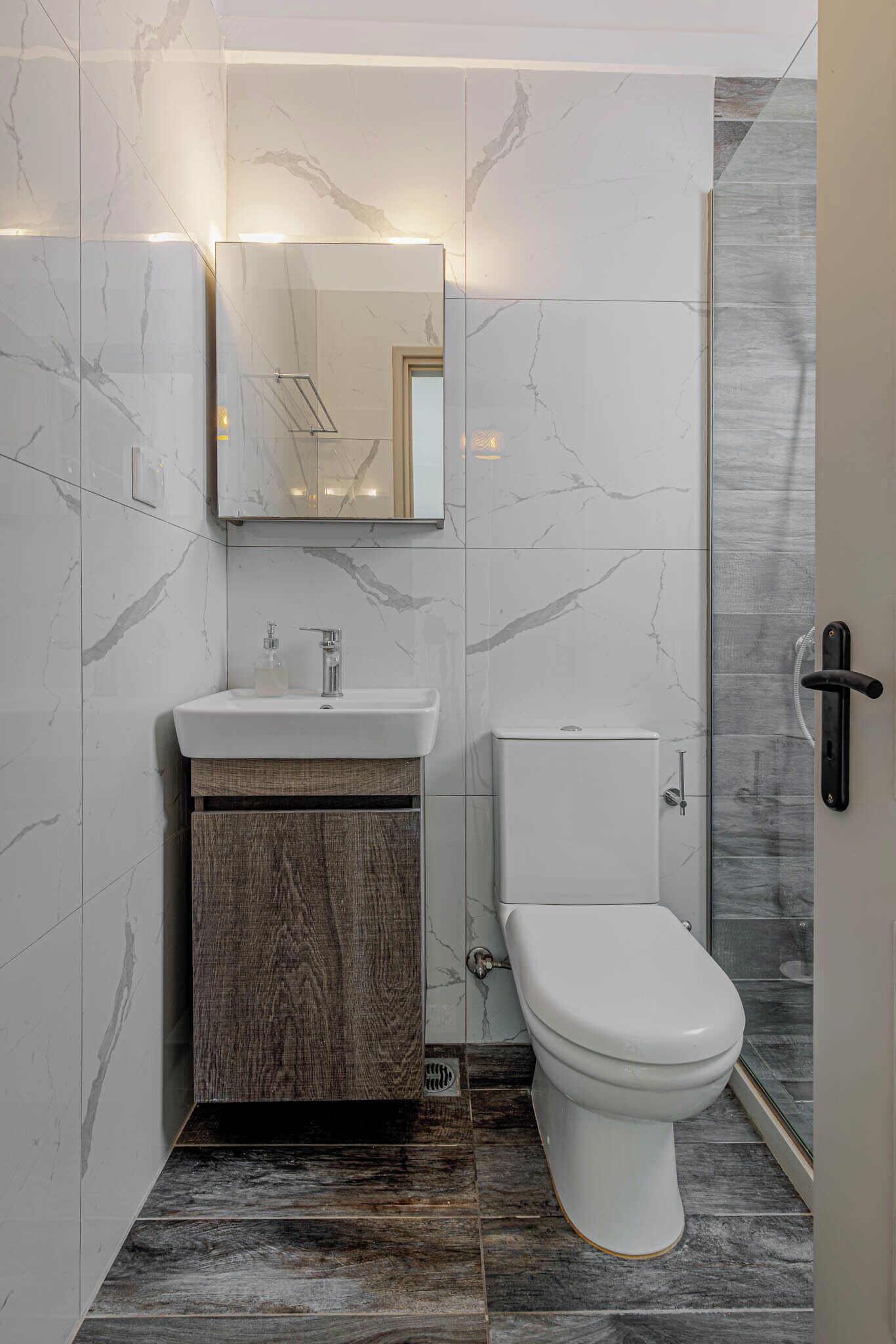 Un baño con pared de mármol, un lavamanos blanco, un mueble de madera, un inodoro blanco y una mampara de cristal para la ducha.