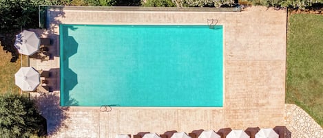 Vue aérienne d’une piscine rectangulaire entourée d’une terrasse en bois. On y voit plusieurs parasols blancs et des chaises longues disposés le long de la terrasse.