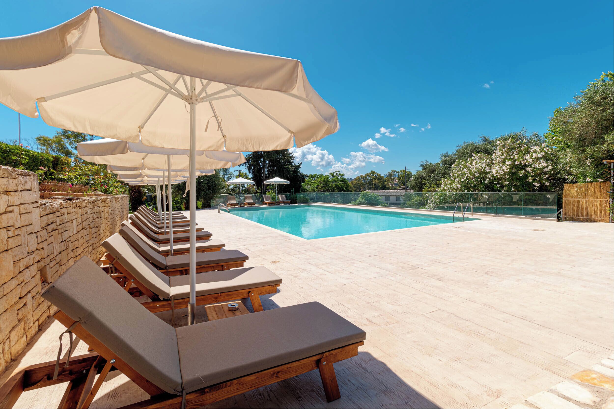 A poolside area with lounge chairs and umbrellas, a stone wall, and a clear blue sky.