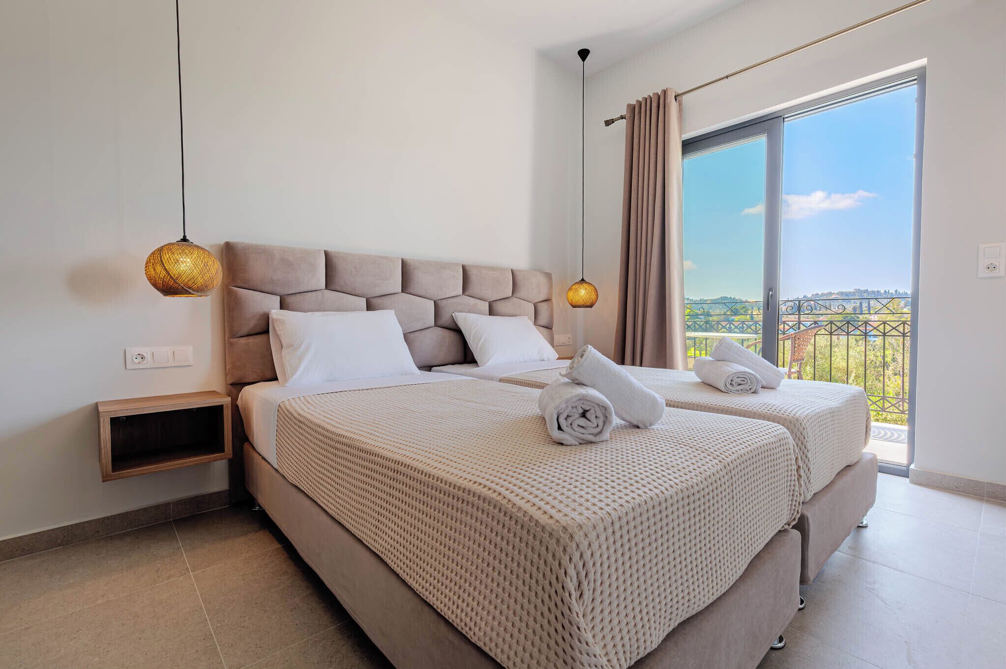 A bedroom with two beds, a nightstand, a hanging lamp, and a balcony with a view.