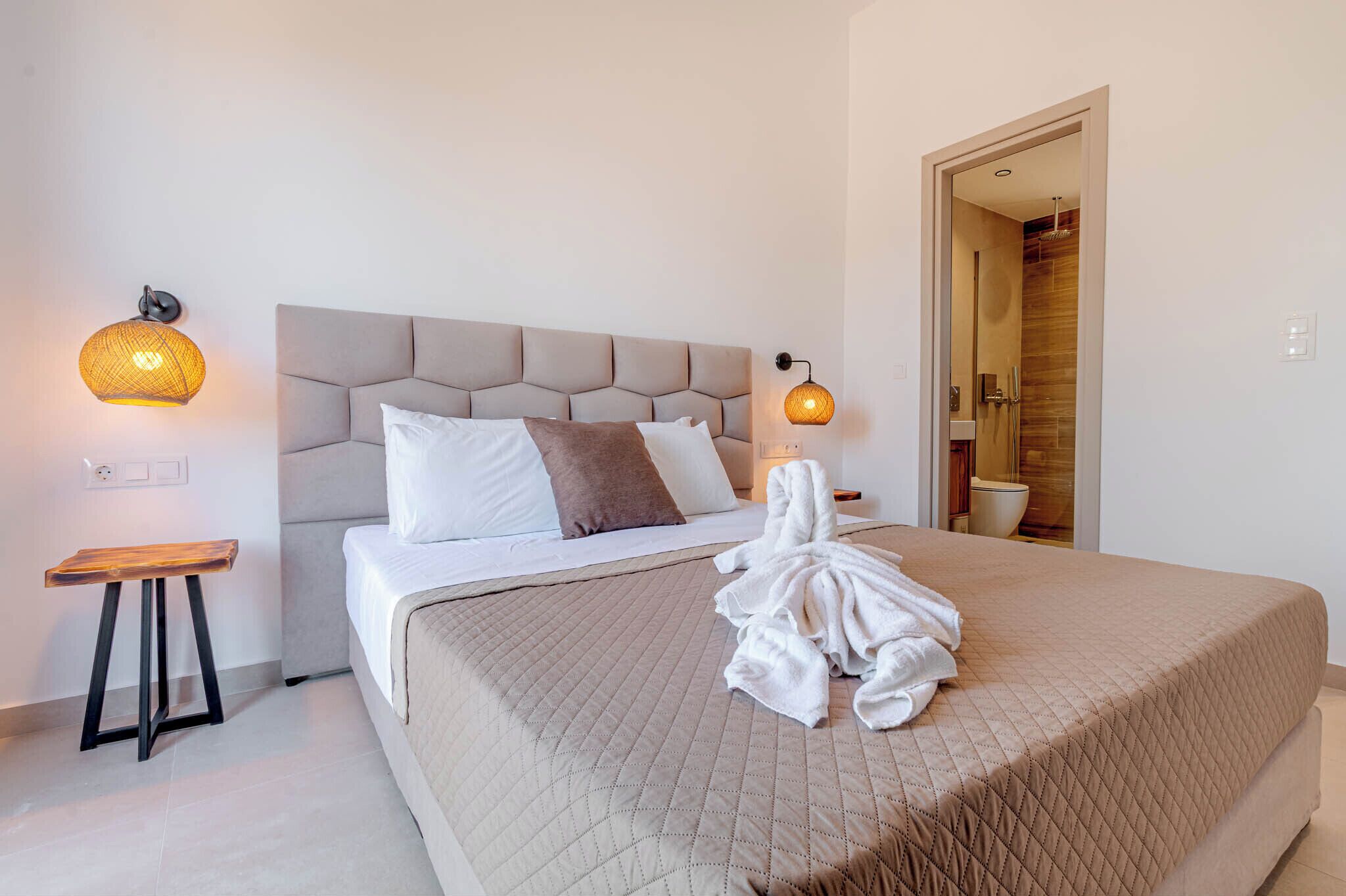 Habitación de hotel con cama, dos lámparas fijadas a la pared, un taburete de madera y un baño visible a través de una puerta abierta.