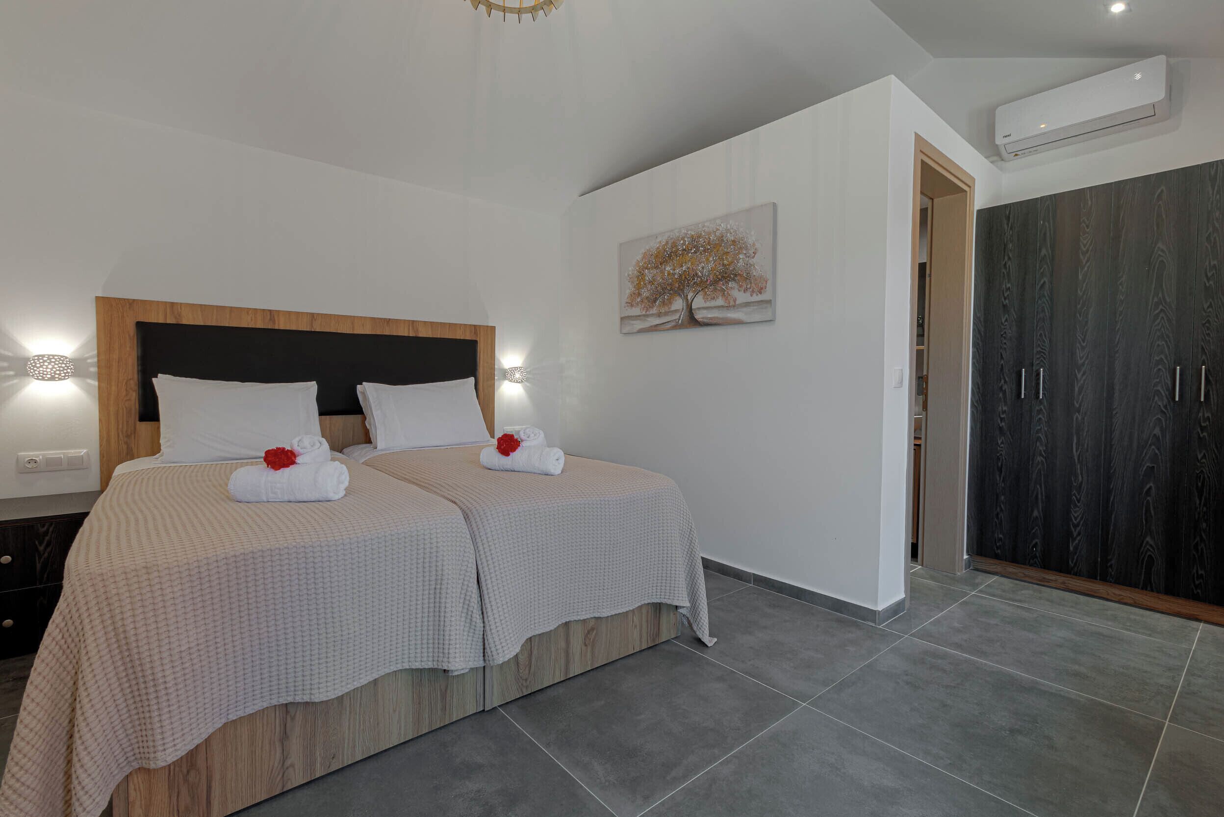 Un lit double avec deux jeux de draps blancs et une fleur rouge, une œuvre d’art représentant un arbre accrochée au mur, et une armoire aux panneaux en bois foncé.