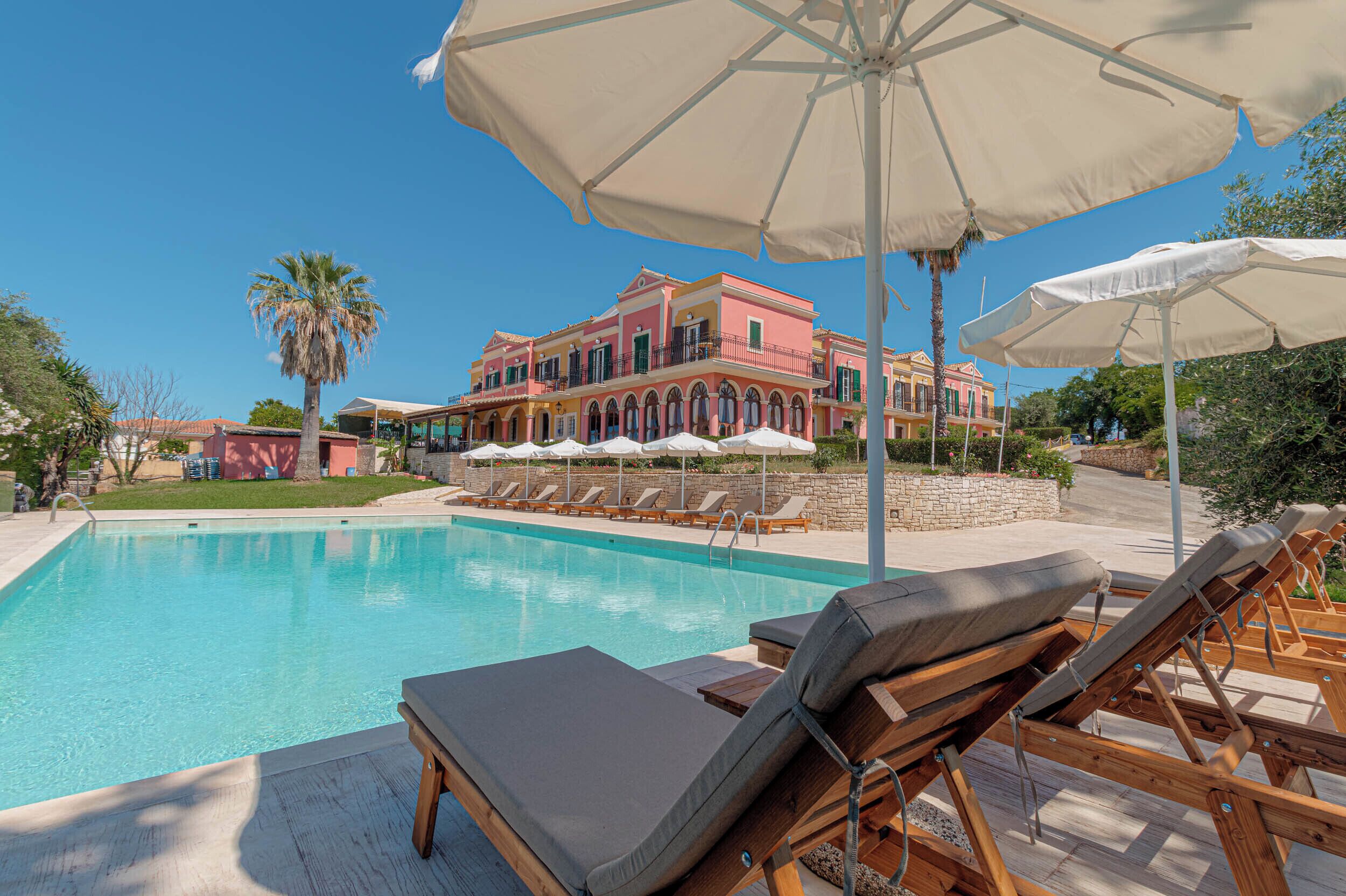 A pool area with lounge chairs and umbrellas, a large building with balconies in the background, and a clear blue sky.