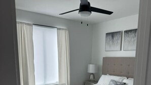 Ein Schlafzimmer mit einem Bett, einem Deckenventilator, einem Nachttisch mit Lampe und zwei abstrakten Bildern an der Wand.