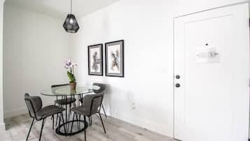 A dining area with a glass table and four chairs, a potted plant, and two framed artworks on the wall.