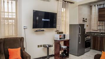 A living room with a flat-screen TV, a sofa, a coffee table, and a kitchen area with a refrigerator and stove.