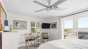 A bedroom with a bed, two chairs, a desk, a ceiling fan, a TV, and a balcony with a view.