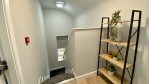 Una escalera estrecha con piso de madera, una alfombra pequeña, barandilla de metal y un estante con plantas.