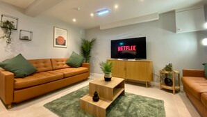 Una sala de estar moderna con un televisor de pantalla plana grande que muestra 'NETFLIX', un sofá de cuero marrón, una mesa de centro de madera y una alfombra verde.