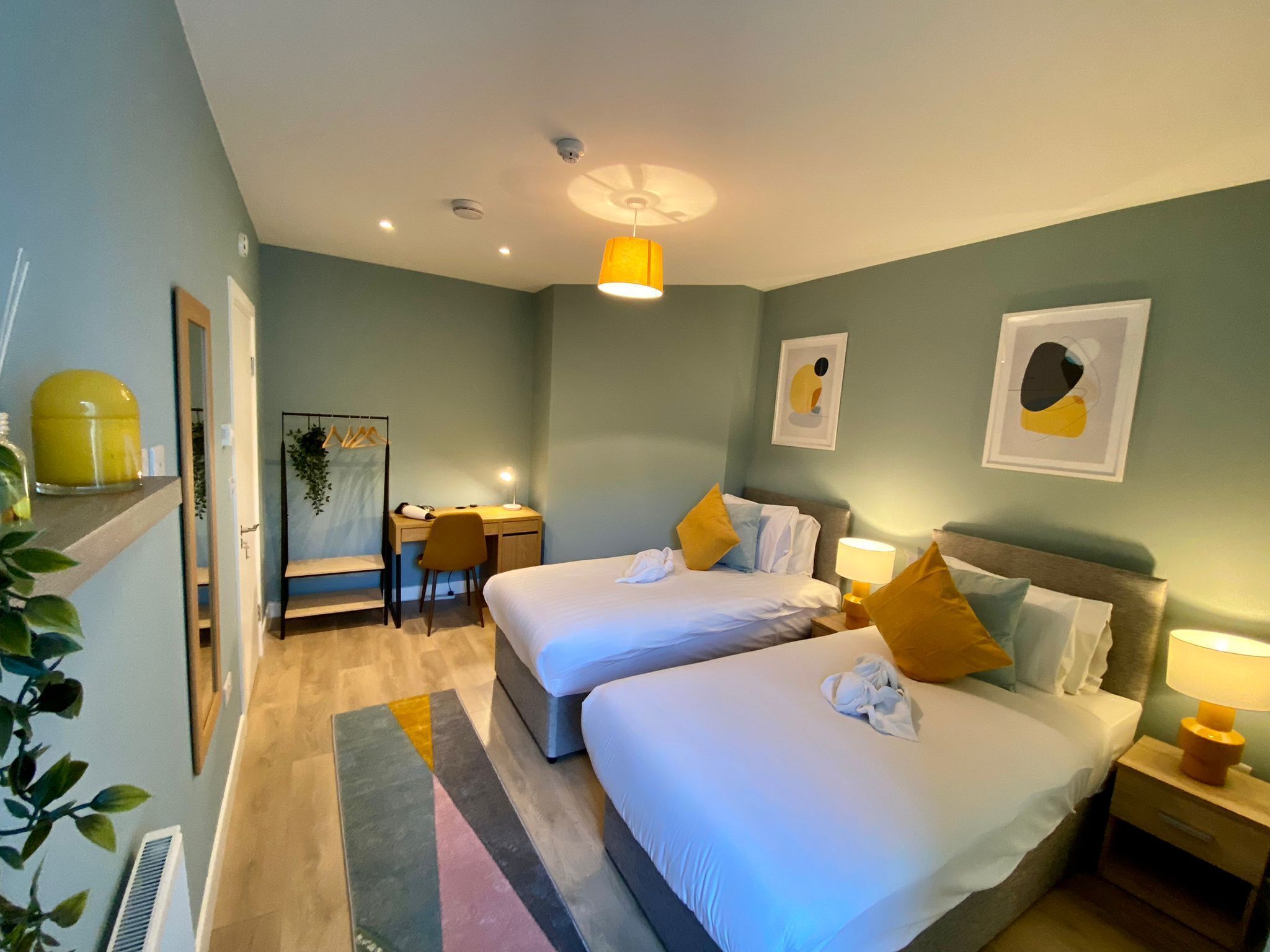 A hotel room with two beds, a desk, a chair, a lamp, and artwork on the walls.